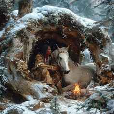 a person sitting next to a white horse in the snow near a fire pit and trees