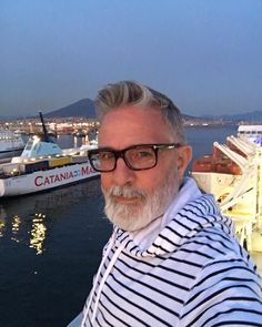 a man wearing glasses and a striped shirt standing in front of a harbor with boats