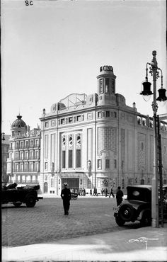 an old black and white photo of people walking in front of a building with cars parked on the street