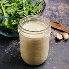 a glass jar filled with dressing next to a bowl of greens and garlic on a table