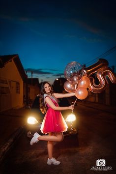 a woman in a red dress is holding balloons and standing next to a black car