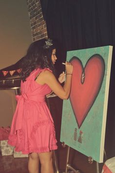 a woman in a pink dress is painting a heart on a canvas with a brush