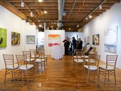 people are standing in an art gallery looking at paintings on the walls and chairs around them