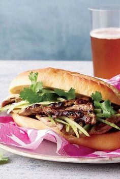 a sandwich with meat, lettuce and cilantro on a plate next to a glass of beer