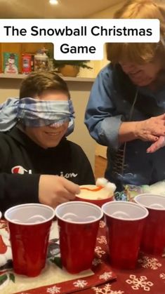 two people sitting at a table with cups on it and one person wearing blindfolds