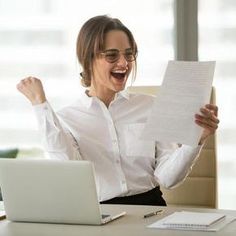 a woman sitting at a desk with her arms in the air and holding a piece of paper