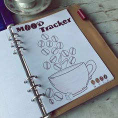 a notebook with coffee beans on it next to a cup of coffee and saucer