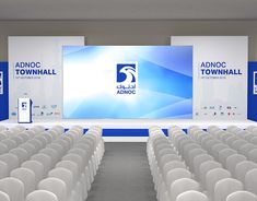 rows of white chairs in front of a large screen with an adnoc town hall logo on it