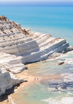 people are walking on the beach near white cliffs and blue water in the distance, with one person standing at the edge of the cliff