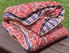 a red and blue blanket sitting on top of a wooden bench in the grass next to a bag