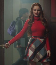 a woman standing in front of a door wearing a red top and plaid skirt with her hands on the door handle