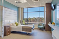 a hospital room with a bed, desk and television on the wall in front of large windows