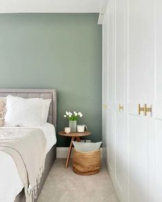 The image shows a bedroom with a sage green wall behind a grey, upholstered bed, dressed with white bedlinen and a grey blanket. To the right of the bed there are floor to ceiling built in wardrobes with white painted doors and brass handles. Dulux Sage Green Bedroom, Mint Room Ideas, Light Grey Green Bedroom, Dulux Pale Sage, Bedroom Color Ideas Green, Wall Colours Ideas Bedroom, Green 09 Lick Paint, Dulux Green Bedroom Wall Colours, Light Green Bedroom Paint