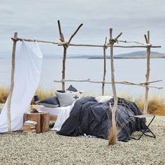 a bed sitting on top of a gravel field next to a body of water under a wooden frame