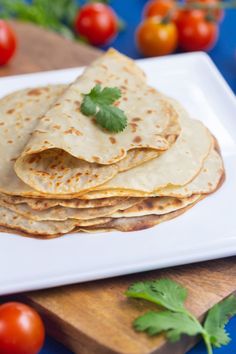 a white plate topped with tortillas on top of a wooden cutting board next to tomatoes