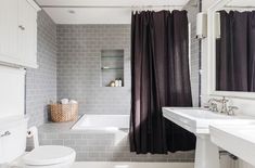 a white toilet sitting next to a bath tub under a bathroom window covered in black curtains