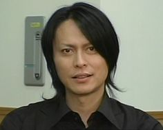 a man with black hair wearing a suit and tie in front of a white wall