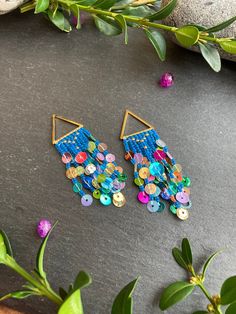 two pairs of earrings with colorful buttons on them sitting next to some leaves and flowers