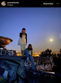 two women standing on top of a stone wall at night with the city lights in the background