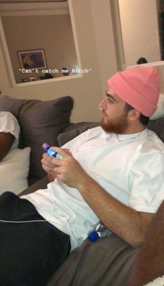 two men sitting on a couch one is holding a remote control and the other has a pink hat