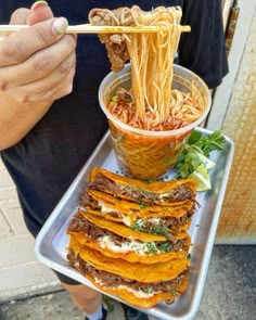 a person is holding a tray with noodles and meats on it while eating from chopsticks