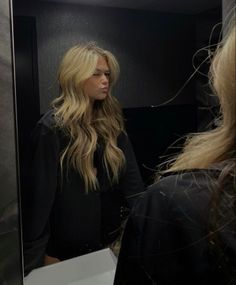 a woman standing in front of a mirror looking at her long blonde hair and wearing a black top