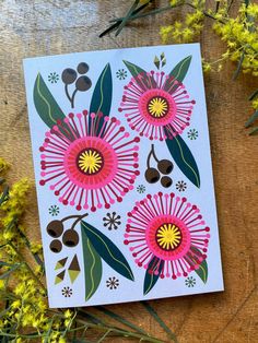 a card with flowers on it sitting next to some green leaves and yellow flowers in the background