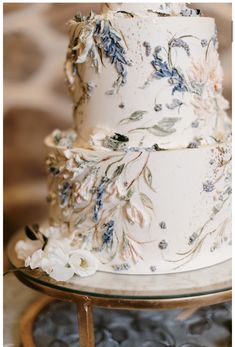 a three tiered wedding cake with blue and white flowers on the top, sitting on a glass stand