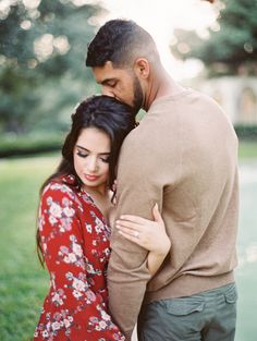 a man and woman standing next to each other in front of a tree with their arms around each other