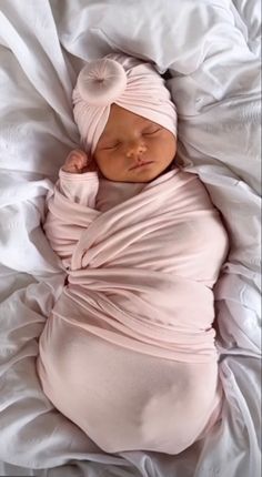 a baby wrapped in a pink wrap sleeping on top of a white blanket and wearing a headband