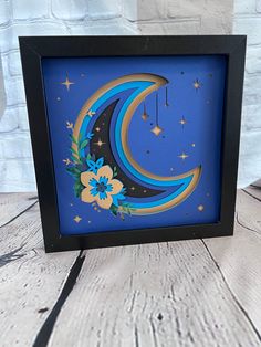 a blue and yellow moon with flowers on it in a black shadow box frame against a white brick wall