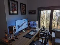 a computer desk with two monitors and speakers in front of a large window overlooking the woods