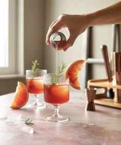 someone is pouring something into two glasses on a table with grapefruits and orange slices