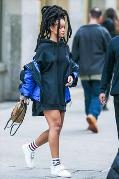 a woman with dreadlocks is walking down the street wearing a black coat and white sneakers