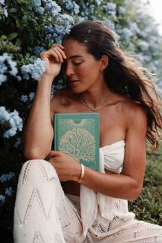 a woman sitting on the ground with a book in her hand and flowers behind her