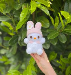 a hand holding up a small crocheted hello kitty doll in front of green leaves