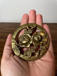 a hand holding a gold brooch with roses on it's center and leaves in the middle