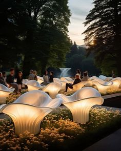 people are sitting on white chairs in the park at night with trees and water behind them