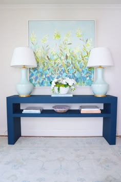 a blue table with two lamps on it and a painting in the corner behind it