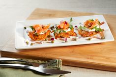 three small appetizers on a white plate next to a knife and fork sitting on a wooden cutting board