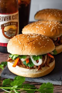 two pulled pork sandwiches sitting on top of a cutting board next to a bottle of beer