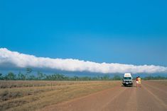 two people standing in front of a van on the side of a dirt road under a cloud filled sky