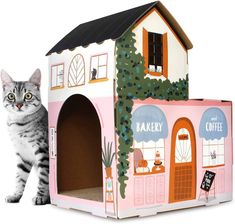 a cat standing next to a pink and white house shaped like a bakery with an awning