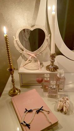 a white vanity topped with a heart shaped mirror next to a pink book and candles