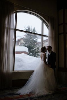 a bride and groom standing in front of an open window looking out at the snow