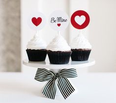 three cupcakes with white frosting and red hearts on top are sitting on a cake plate