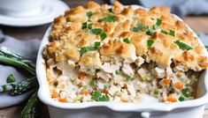 a casserole dish with chicken and vegetables in it sitting on a table next to asparagus