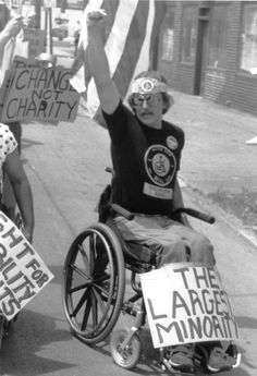 a man in a wheel chair holding up signs while another person holds an american flag