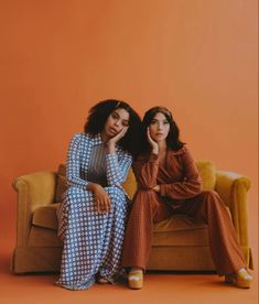 two women sitting on top of a couch next to each other, one in blue and the other in orange