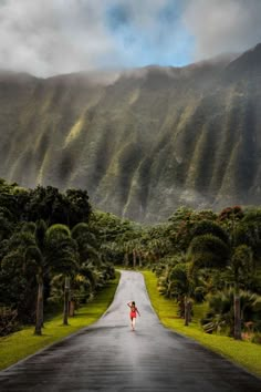 a woman walking down the middle of a road in front of a lush green mountain